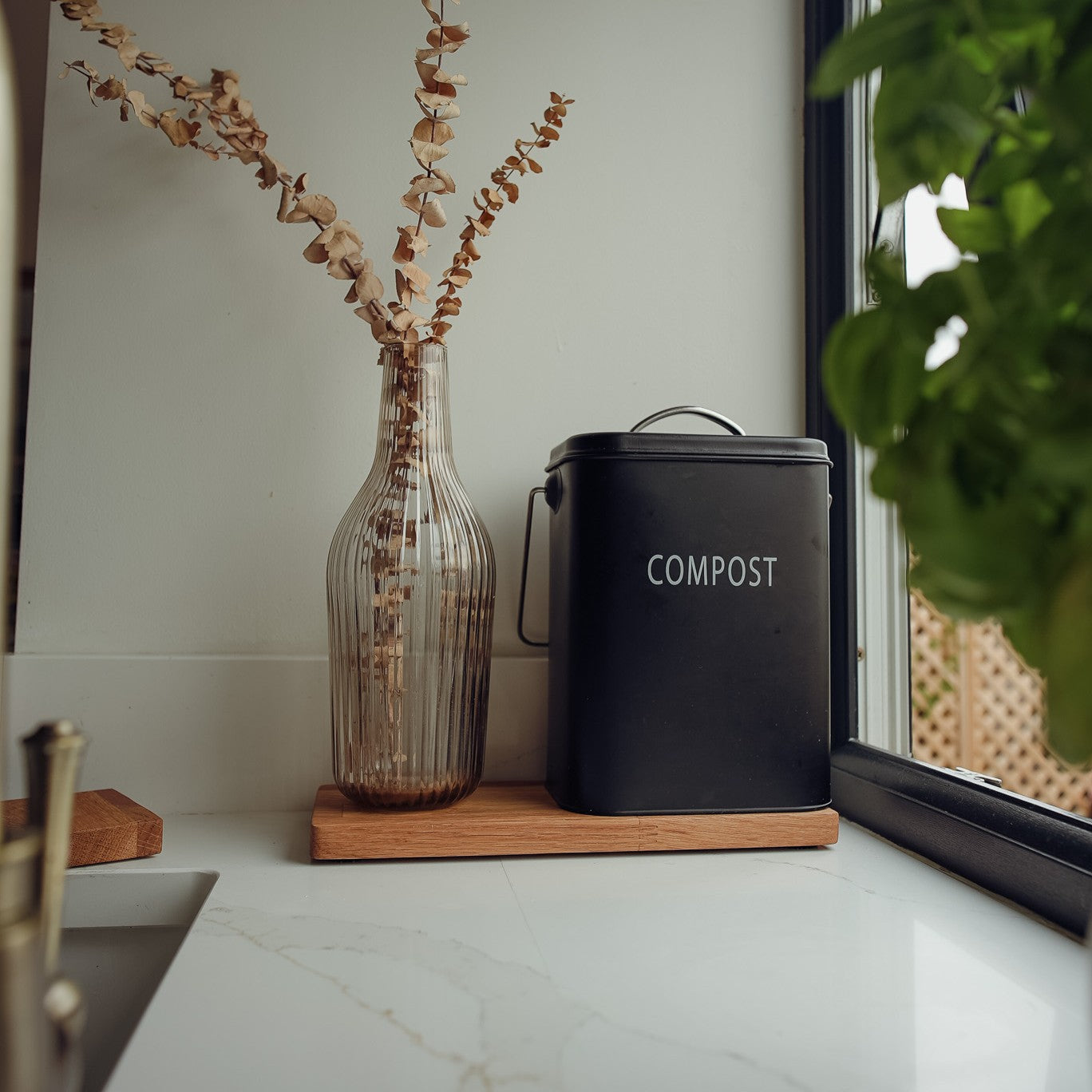 Black compost bin on a kitchen counter sat on a long piece of wood with a vase of dried flowers and a window in the background.