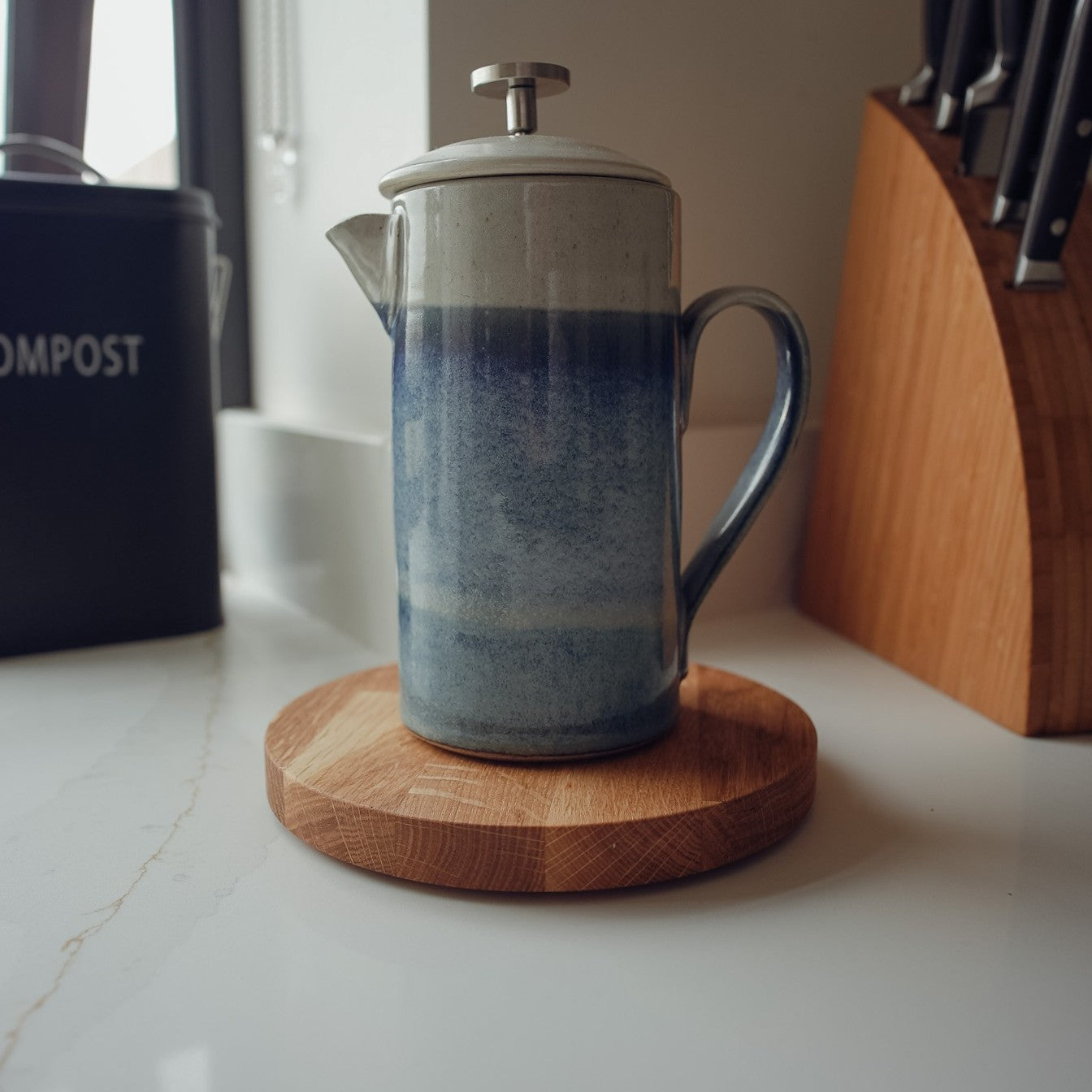 Blue ceramic French press on a large round wooden coaster with a knife block in the background