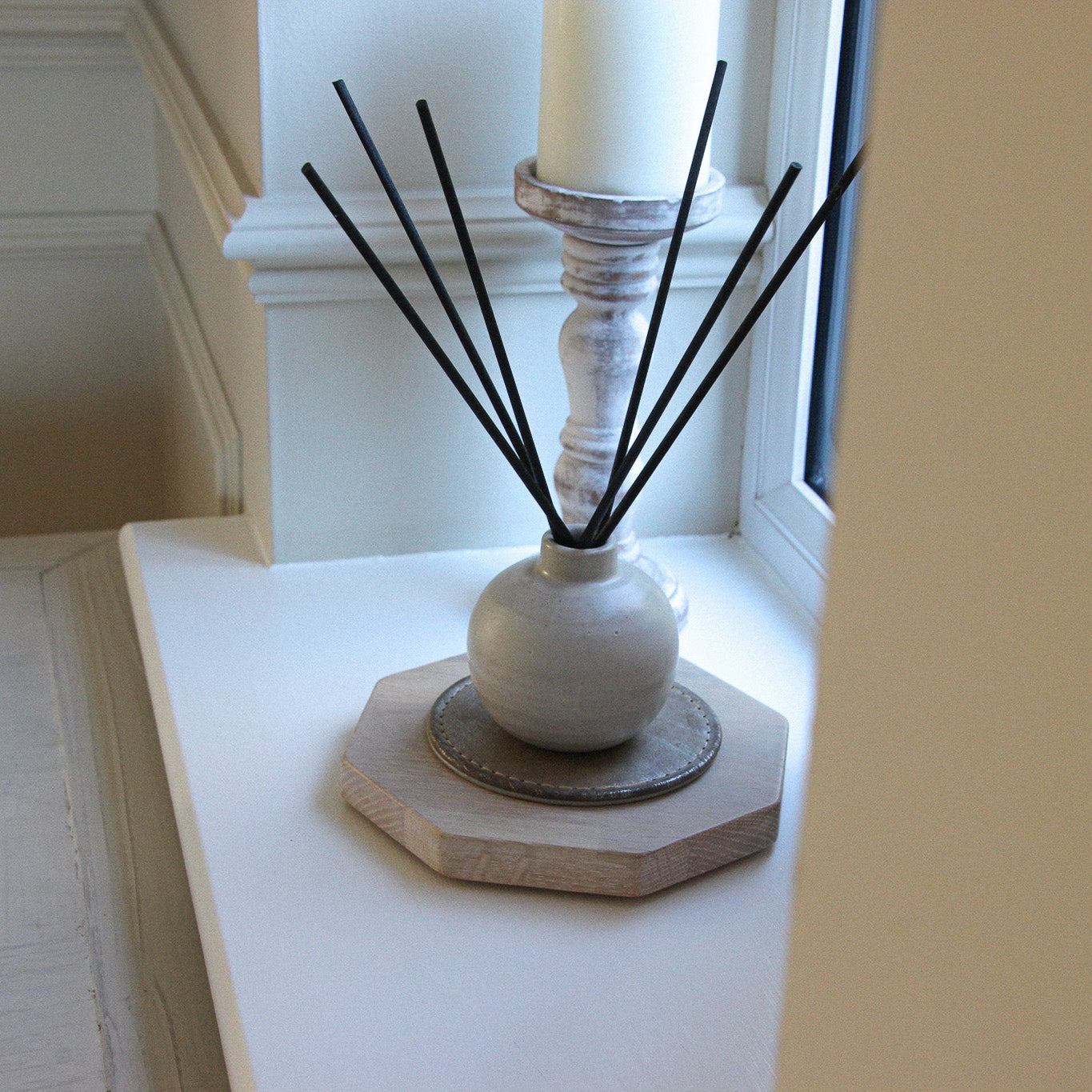Decorative diffuser with black reeds and a white candle on a limed oak wooden 50 pence piece display board.