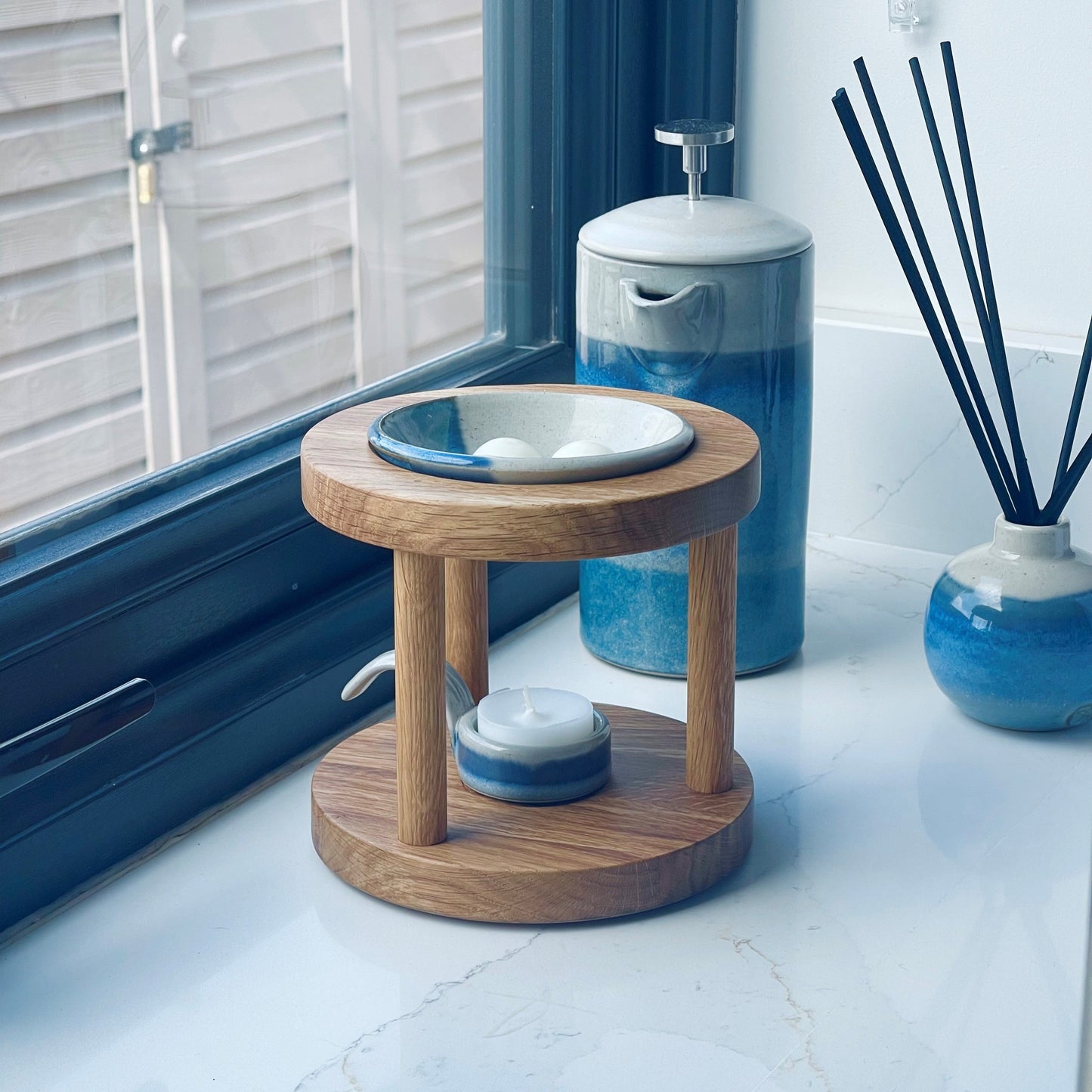 Round shaped wooden wax melt burner in warm oiled oak shade with blue glazed pottery tealight spoon and wax melt dish on a windowsill