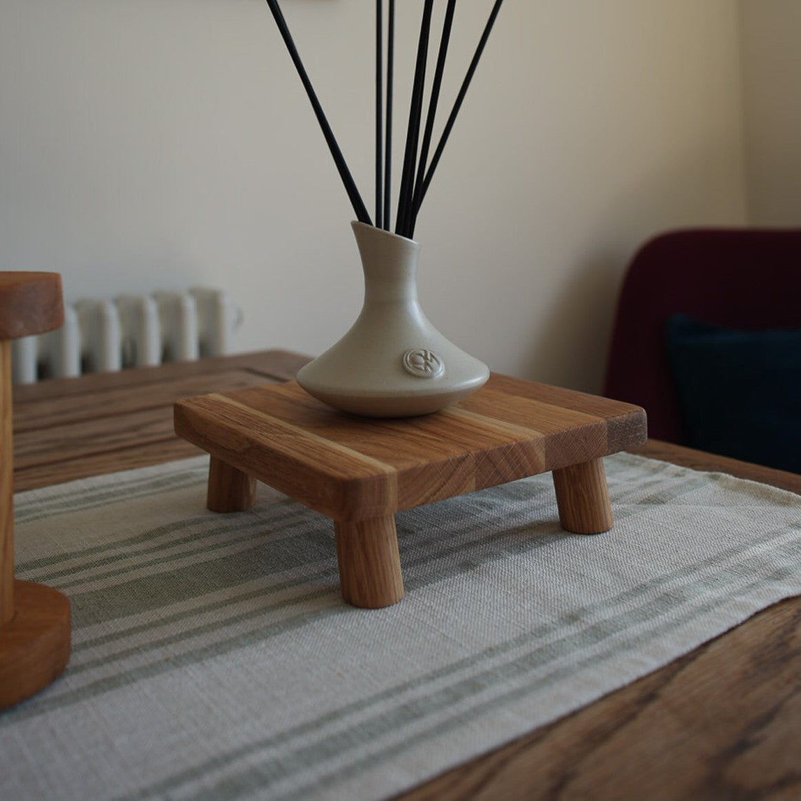 An oiled oak, square plant pot stand topped with a small ceramic reed vase, set on a striped mat atop a wooden table.