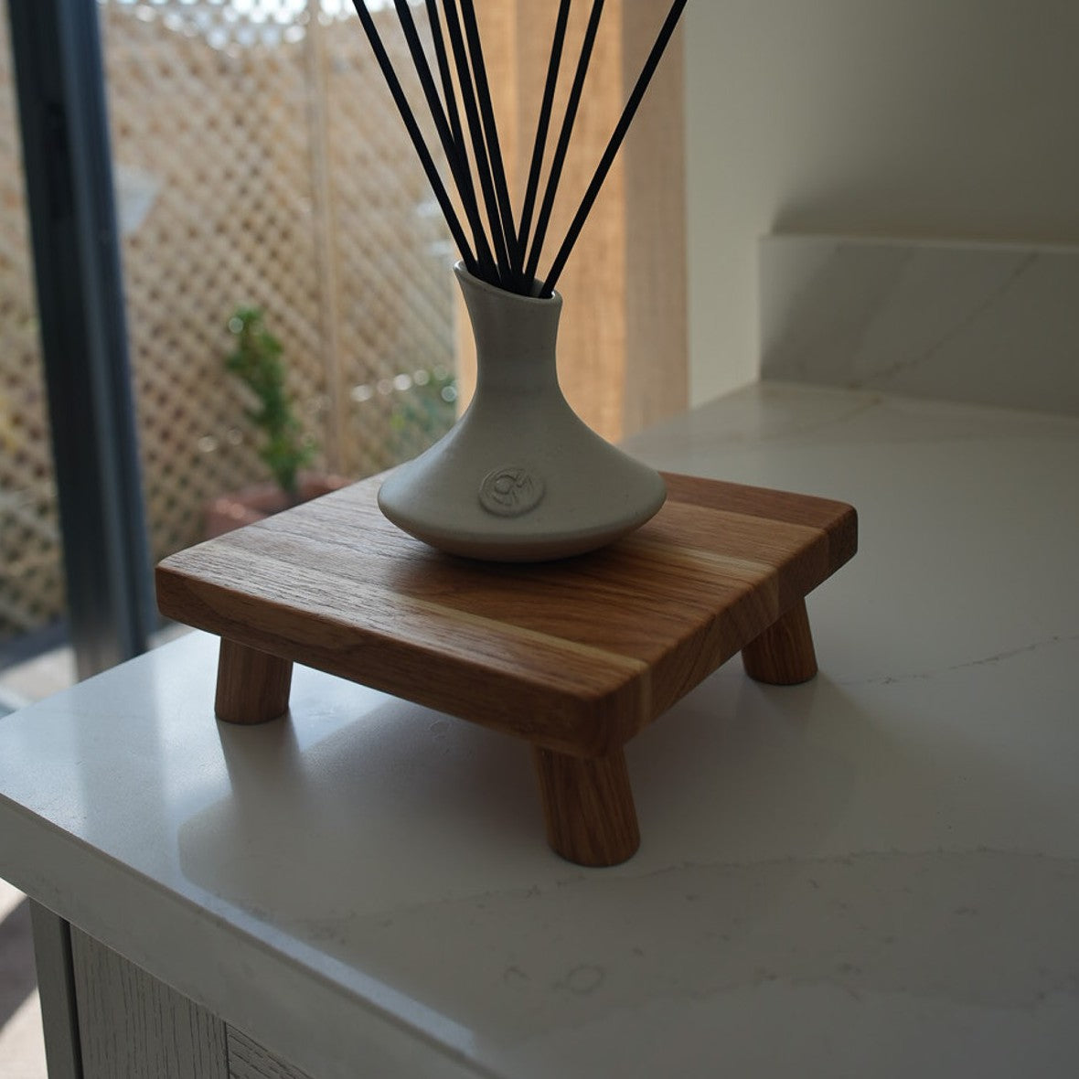 A square-shaped, oiled oak plant pot stand with a small ceramic vase on top containing reeds, placed on a striped mat on a wooden table.