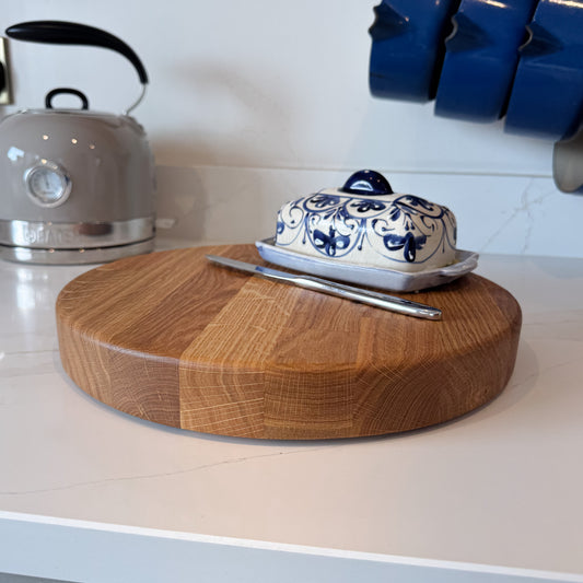 Round wooden oak chopping board placed on white marble kitchen counter with a blue patterned butter dish and silver knife on top with a grey kettle in the background and some hanging blue saucepans.