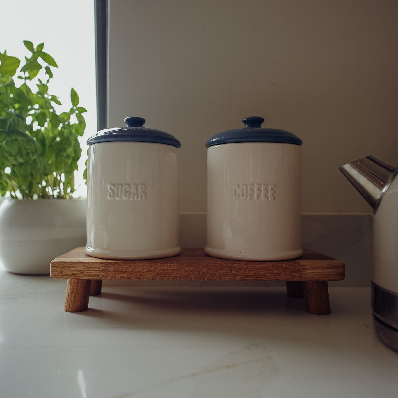 Two ceramic jars labelled 'Sugar' and 'Coffee' on a wooden stand with legs for home styling purposes with a plant and kettle in the background.