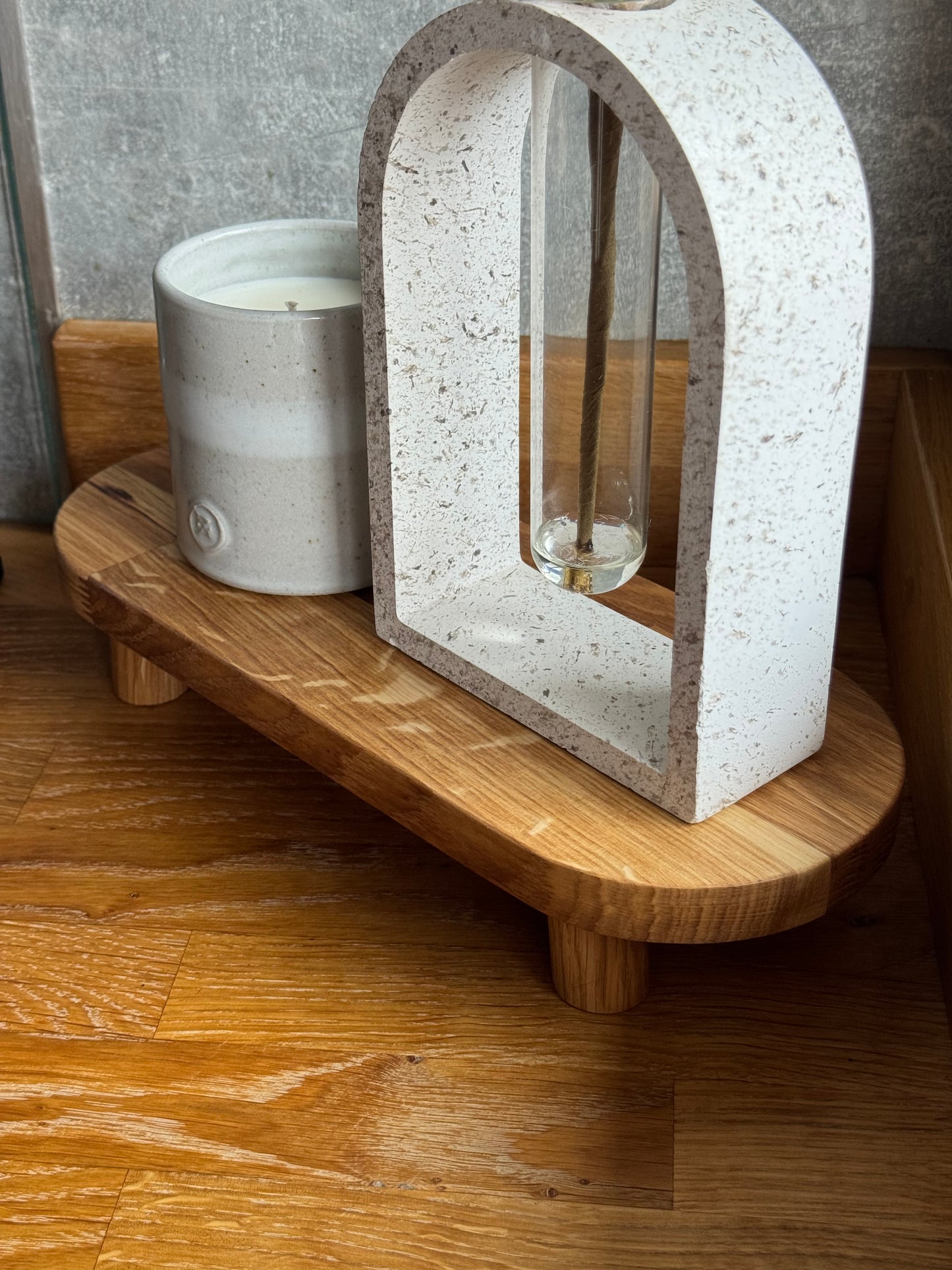 The Alyn Oak Riser is pictures on an oak worktop with a candle and another cement style ornament on top. The riser is oblong with rounded edges and 4 very short feet. You can clearly see the grain in the wood with the oiled oak orange tones