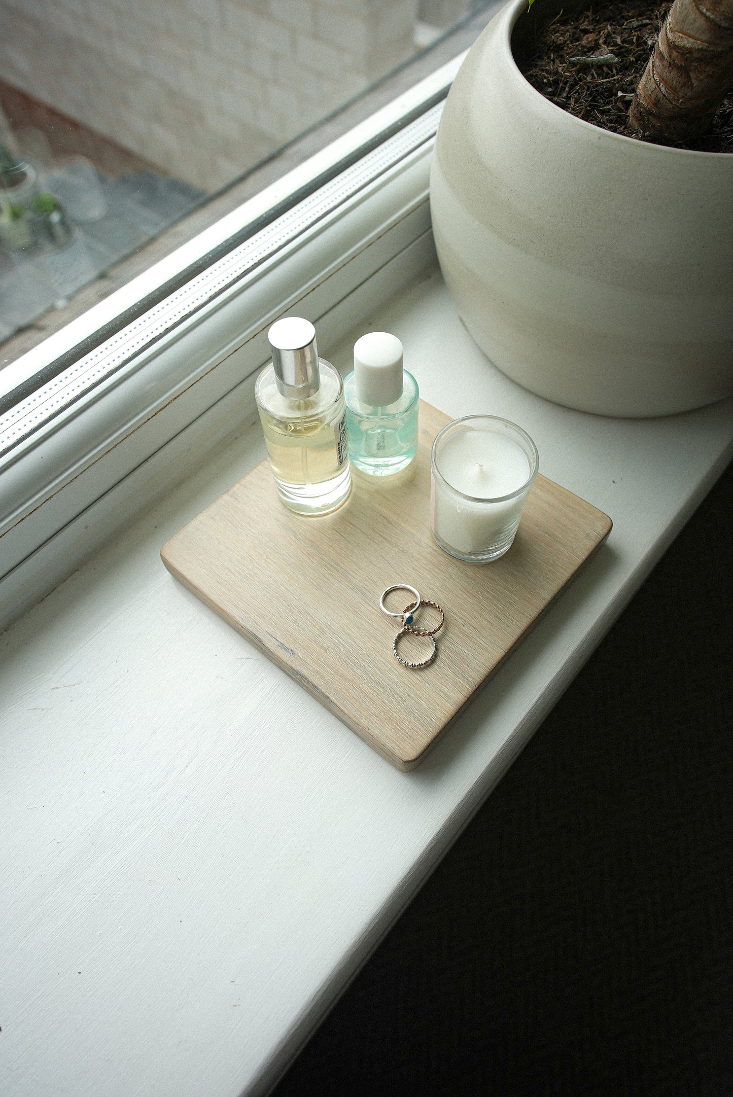 Two bottles of perfume, a candle, and a square wooden coaster made from oak in a light wash shade on a windowsill with a plant in the background. The purpose of this image is to show the display mat and how it can be used for home styling