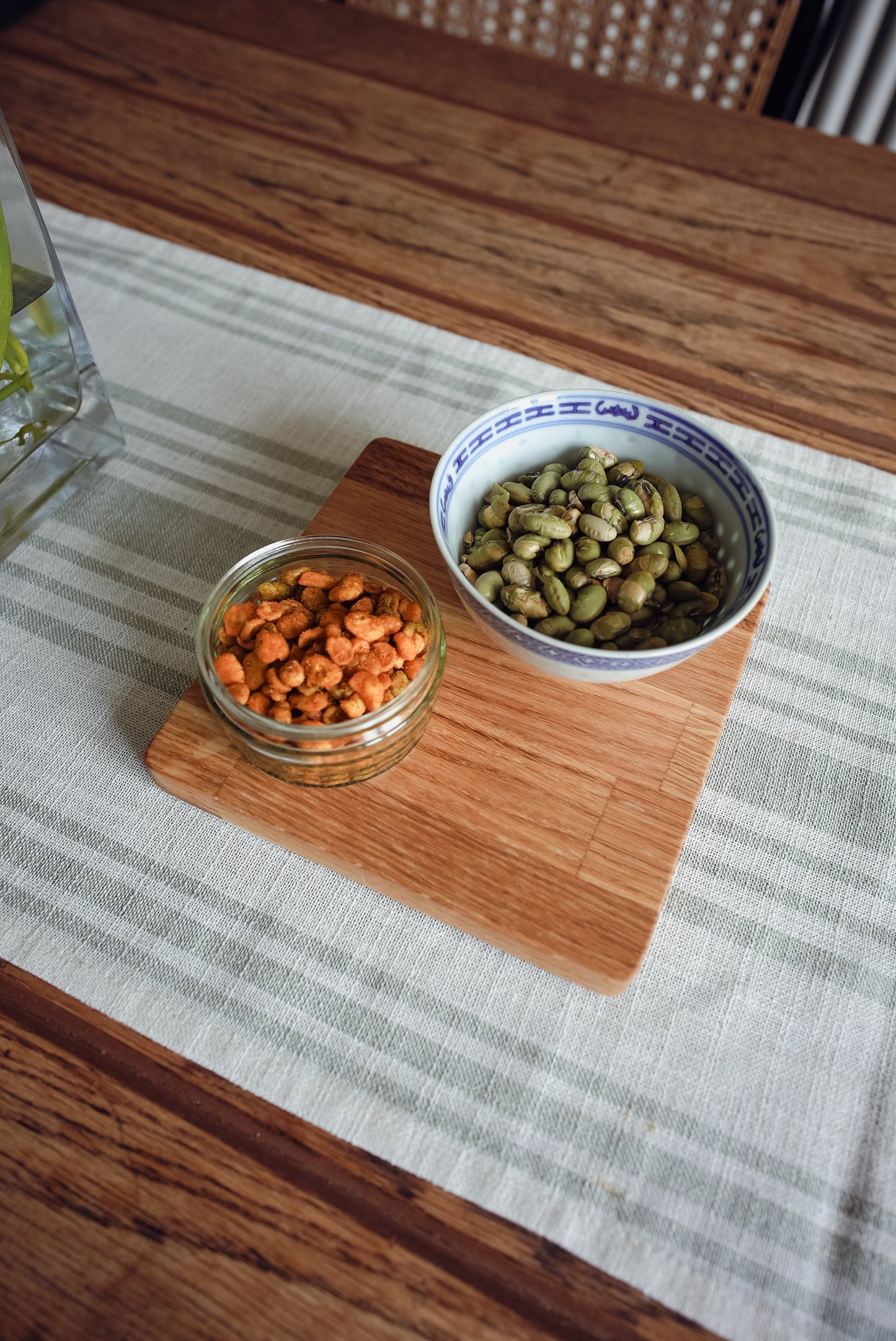 Two containers, one with orange snacks and the other with green beans, on a wooden cutting board/display mat. It is on top of a white and sage stripey runner on an rustic oak table. The mat is finished with an oak oil which brings out the orangey natural grain of the oak.