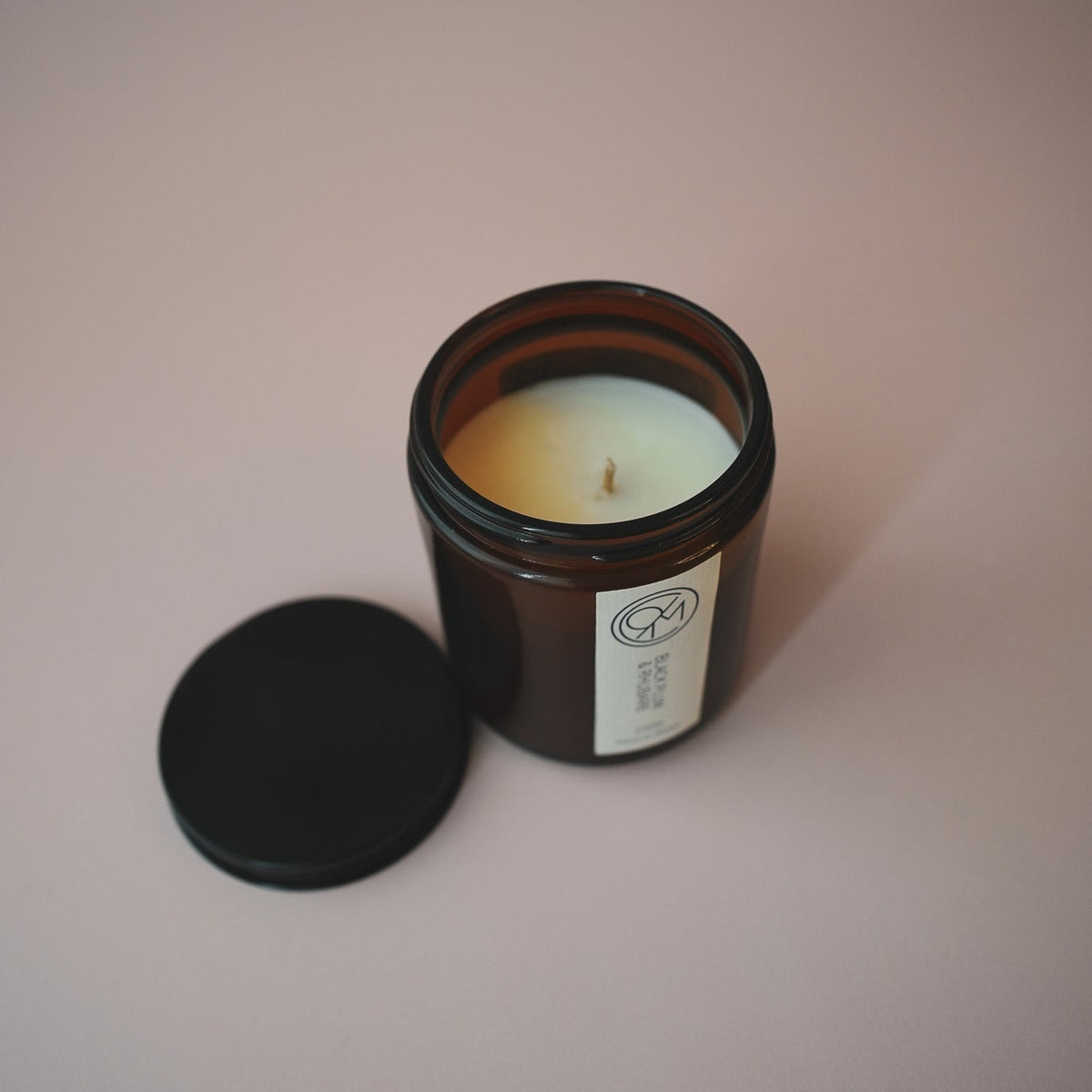 An open amber glass candle jar with cream-colored wax and a black lid resting beside it on a solid, muted dusty pink background.