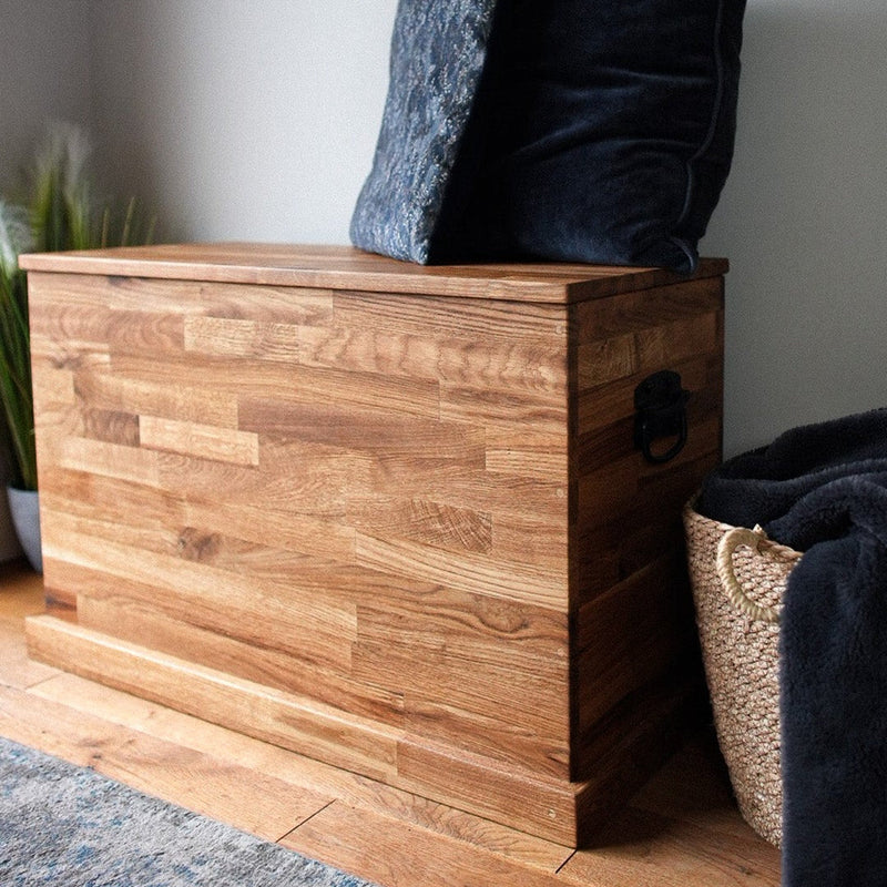 A rustic wooden storage chest with a hinged lid, styled with two dark throw pillows and a navy blanket draped over a woven basket in a home setting.