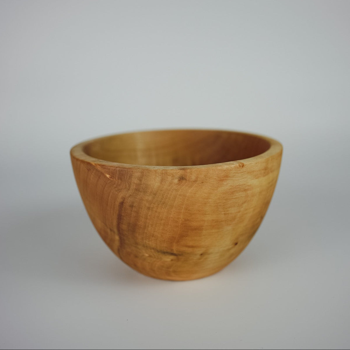 A small, handcrafted wooden bowl with a warm honey-brown finish and visible natural wood grain, set against a plain light gray background.