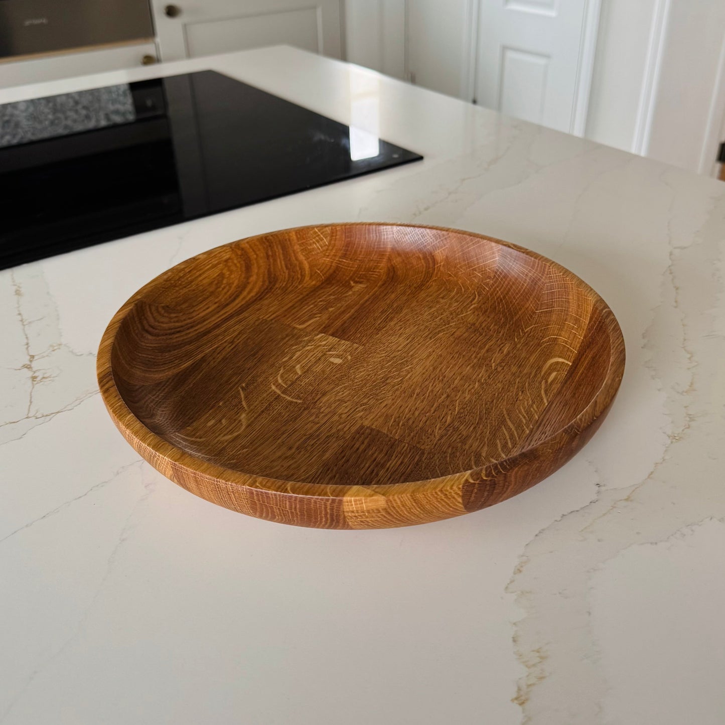 Very large shallow fruit bowl made from engineered oak, the light of the kitchen is reflecting onto the edges of the bowl and you can see the orangey oak grain very well, it's placed on a marble kitchen counter