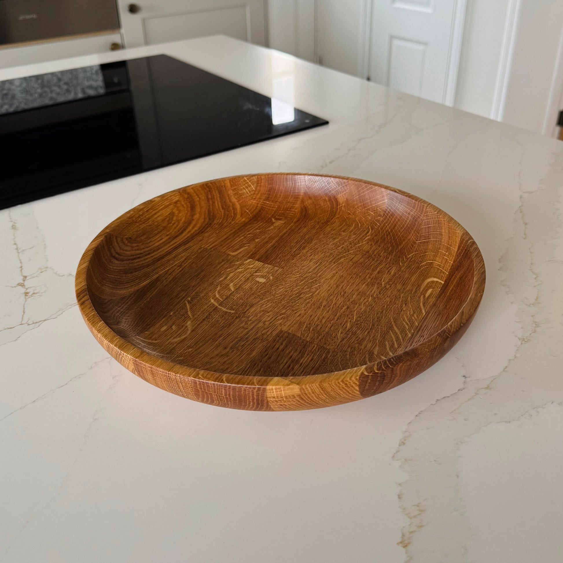Very large shallow fruit bowl made from engineered oak, the light of the kitchen is reflecting onto the edges of the bowl and you can see the orangey oak grain very well, it's placed on a marble kitchen counter