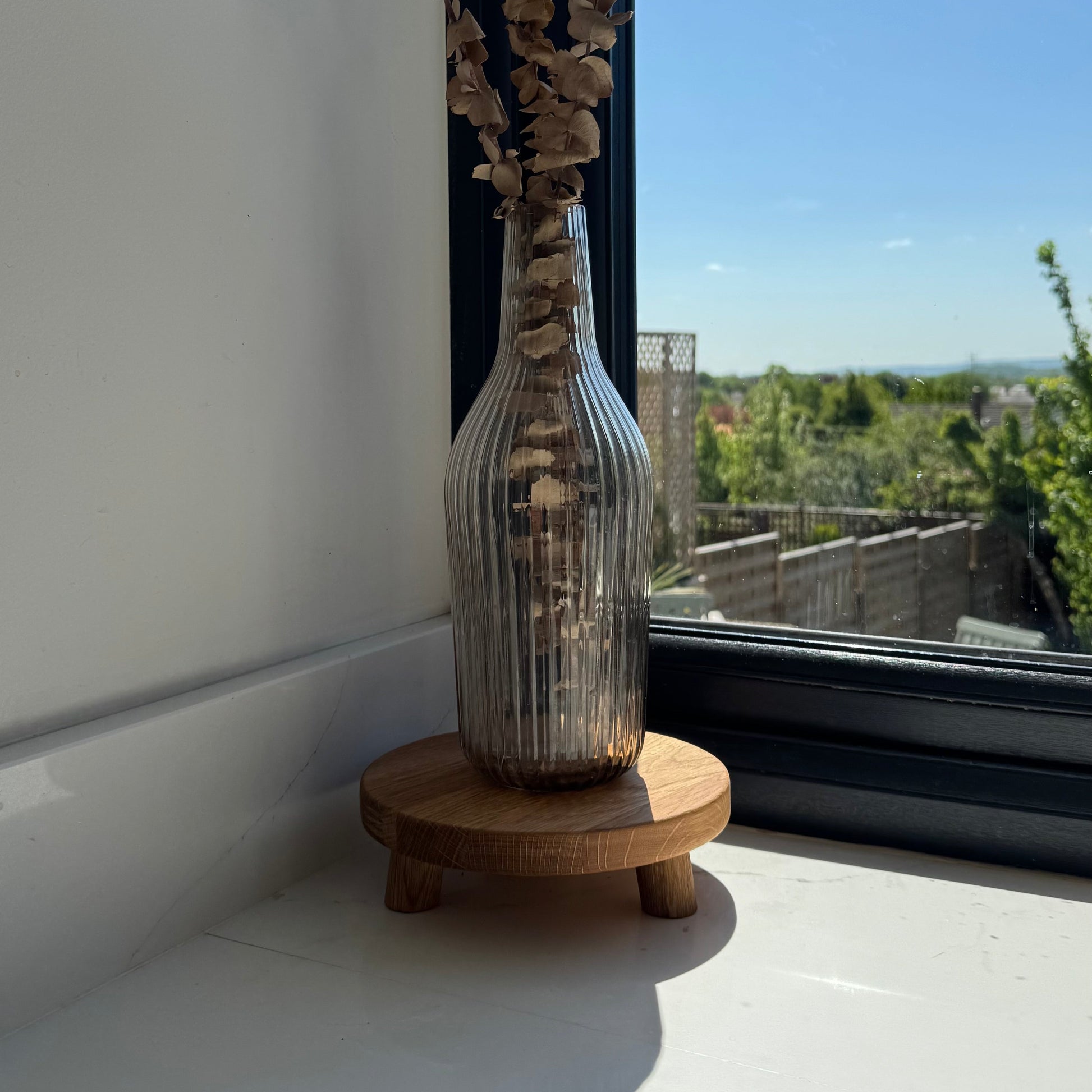 Glass vase with dried plants on a wooden stand with small legs showing the grain of wood by a window with a view of trees and buildings.