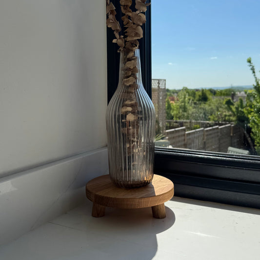 Glass vase with dried plants on a wooden stand with small legs showing the grain of wood by a window with a view of trees and buildings.