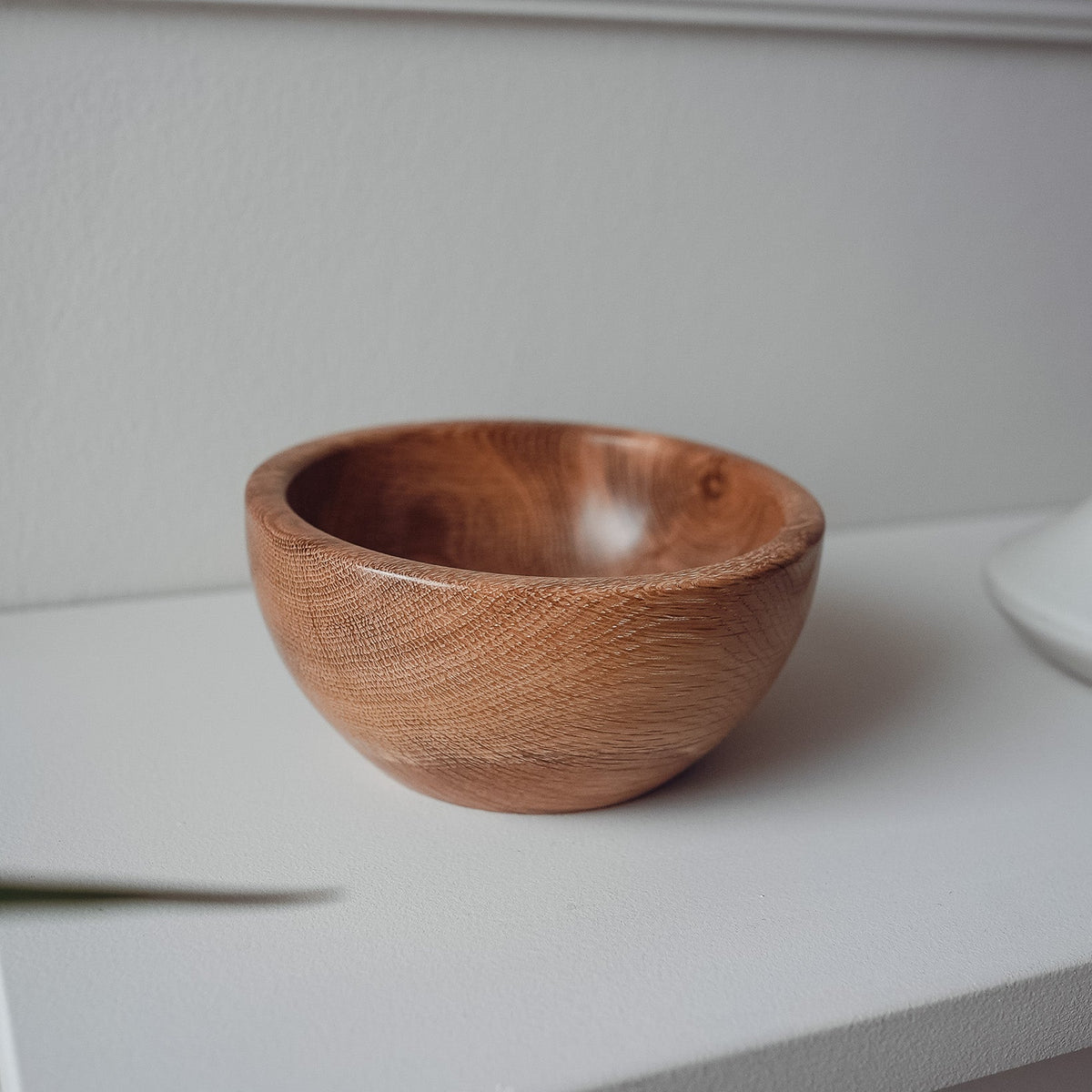 A small handcrafted wooden bowl with visible wood grain, sitting on a white shelf against a white wall.