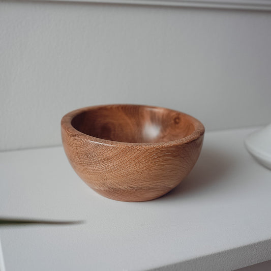 A small handcrafted wooden bowl with visible wood grain, sitting on a white shelf against a white wall.