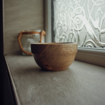A small round wooden bowl with a visible wood grain texture sits on a gray stone or concrete ledge next to a frosted glass window with a leaf pattern.