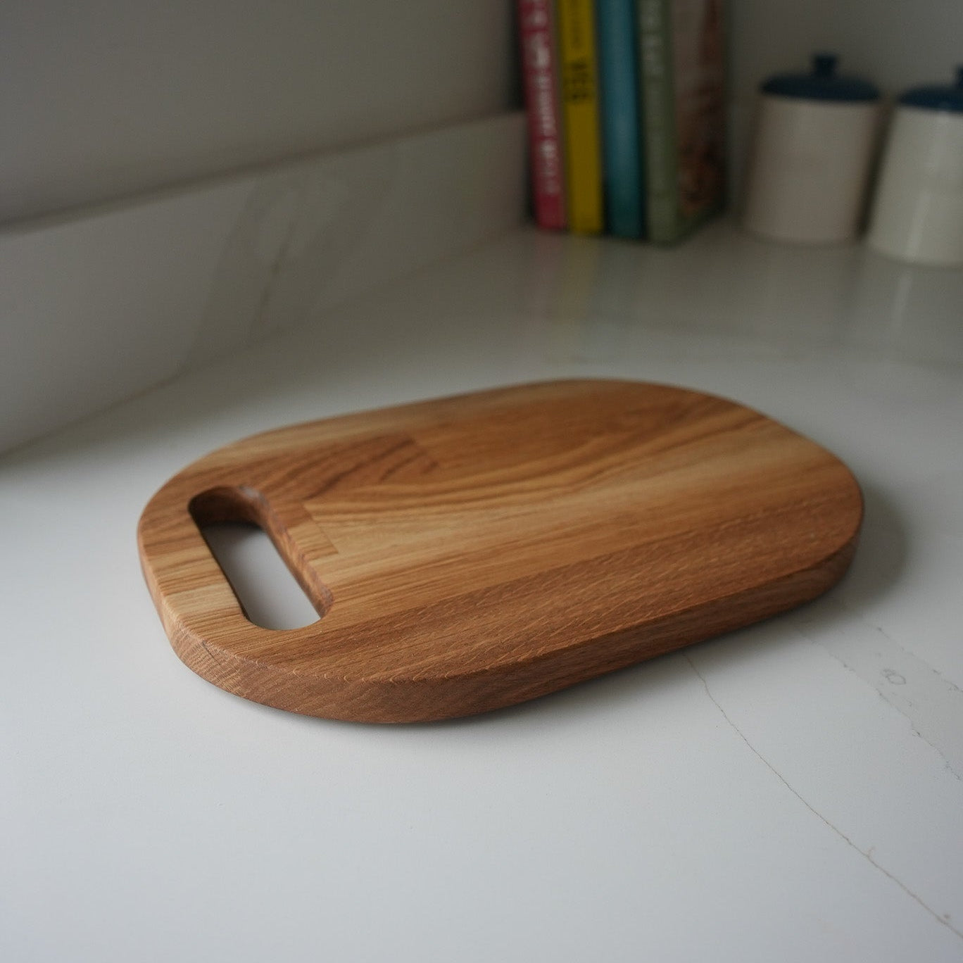 A light brown wooden cutting board with a handle cutout, resting on a white marble countertop with books and canisters blurred in the background.