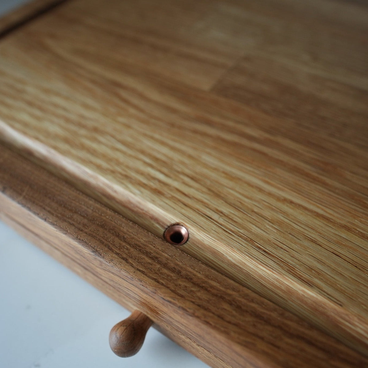 Close-up of a wooden board or tray with a visible tongue-and-groove joint and a small round metal fastener, photographed on a light neutral surface.