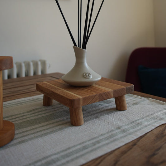 An oiled oak, square plant pot stand topped with a small ceramic reed vase, set on a striped mat atop a wooden table.