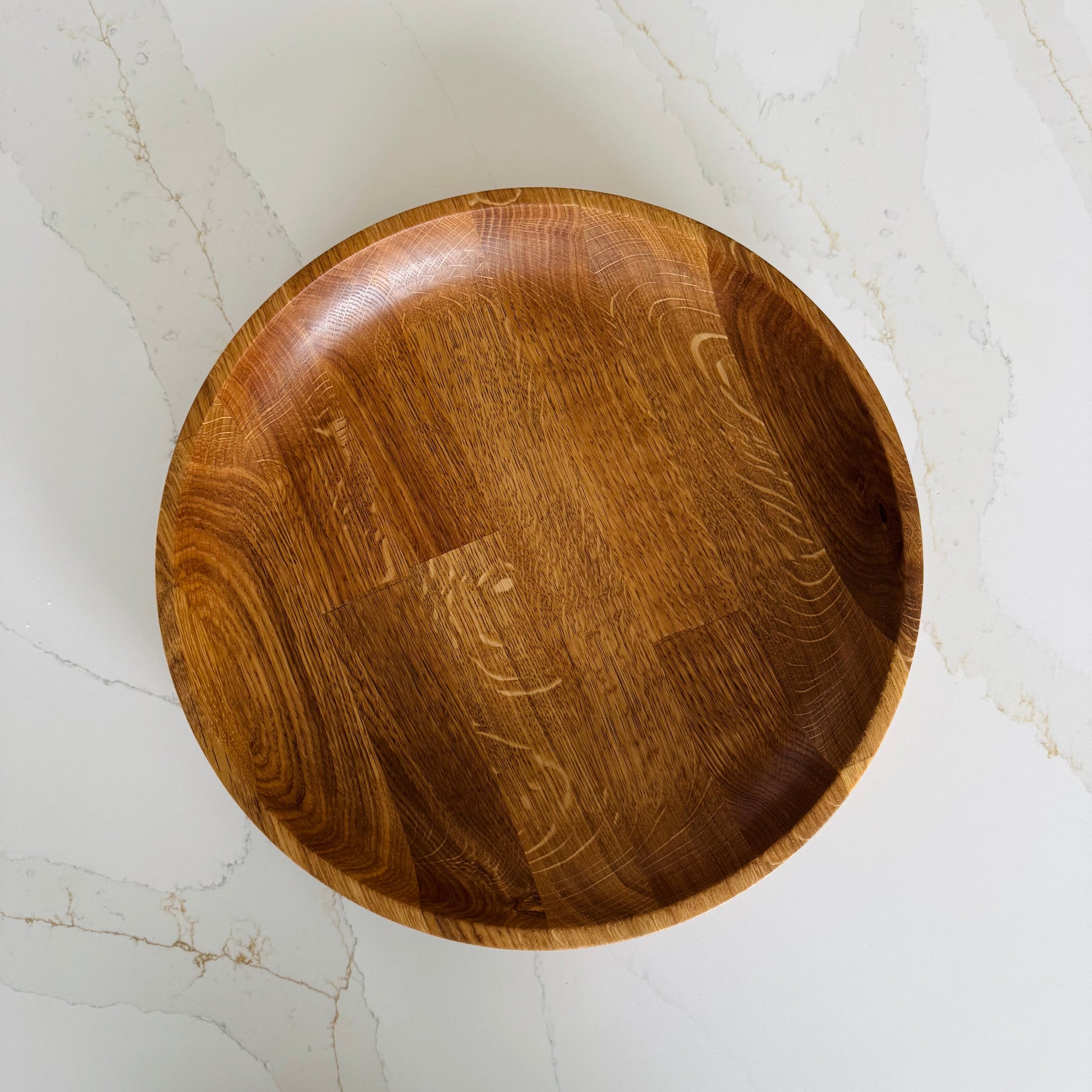 Wooden bowl on a light marble surface, the image is taken from a birds eye view to show the oiled oak finish which shows the natural grain of the wood in an orangey tone.