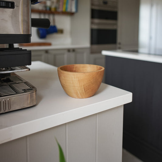 A light brown wooden bowl sits on a white kitchen island counter in a modern kitchen setting with a coffee machine in the foreground and a dark gray cabinet in the background.