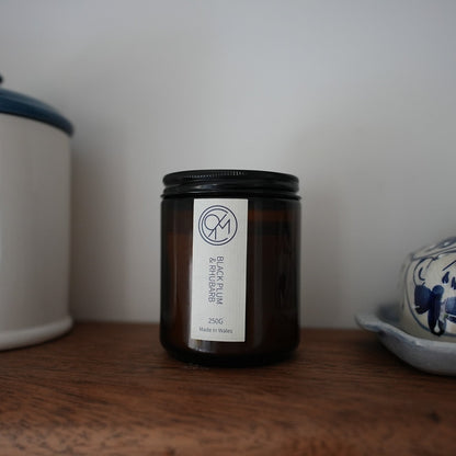 A dark amber glass jar candle with a black lid and a white label reading 'black plum & rhubarb.