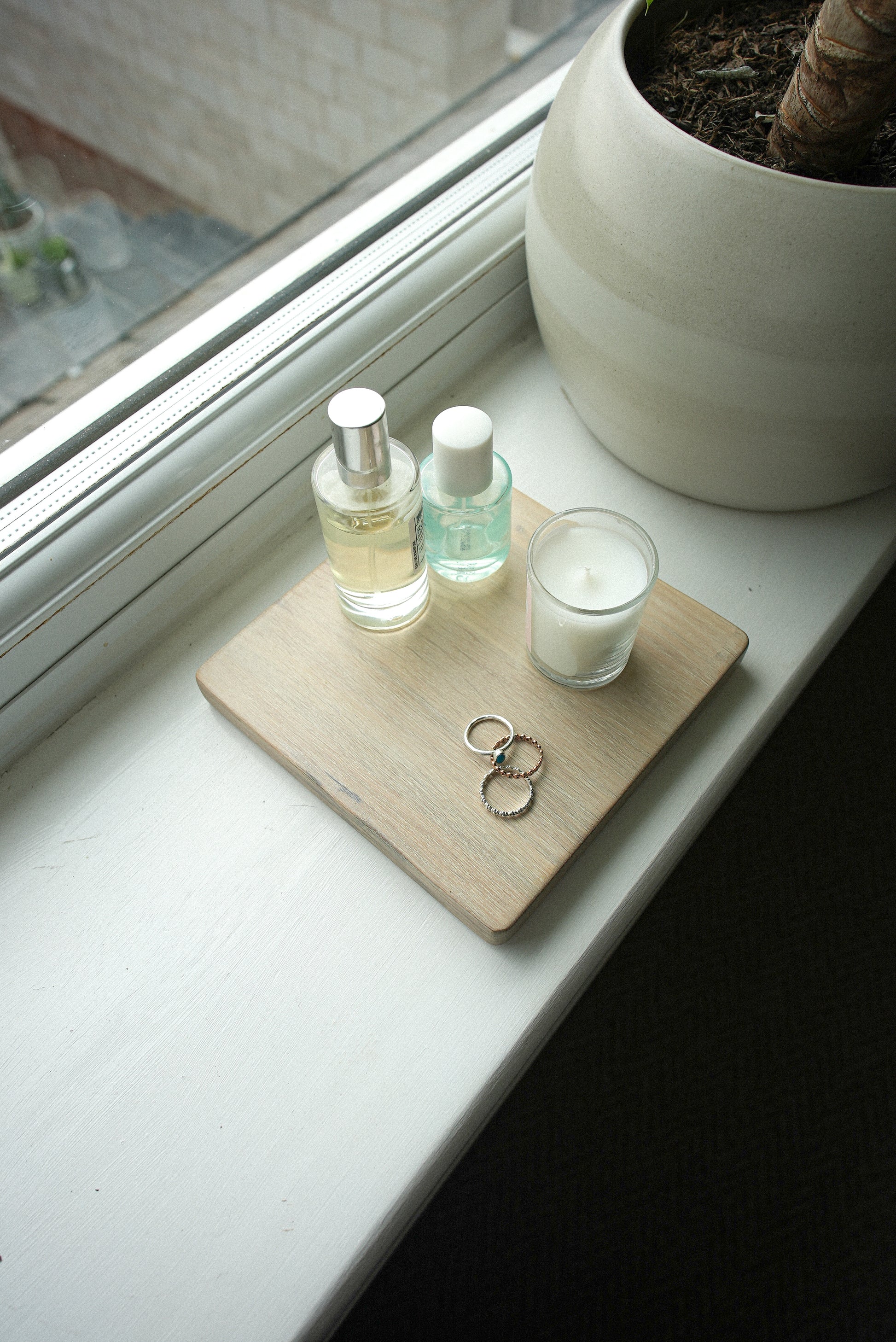 Two bottles of perfume, a candle, and a square wooden coaster made from oak in a light wash shade on a windowsill with a plant in the background. The purpose of this image is to show the display mat and how it can be used for home styling