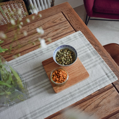 Birdseye shot of an oiled oak display mat placed on top of a white and sage stripey runner on an rustic oak table. The display mat is serving snacks in two bowls. The mat is finished with an oak oil which brings out the natural grain of the oak.