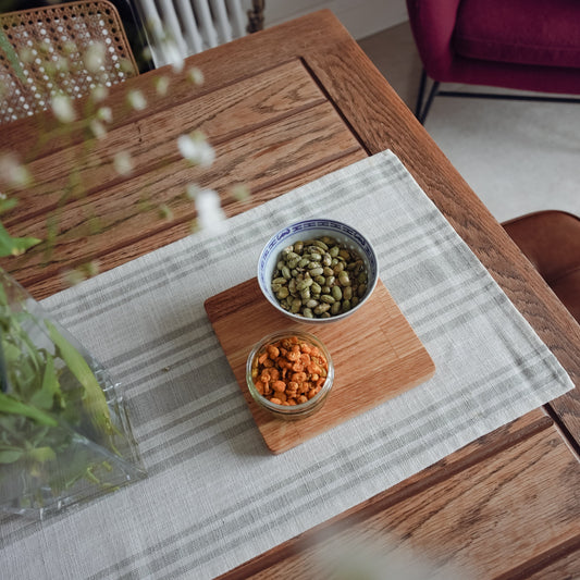 Birdseye shot of an oiled oak display mat placed on top of a white and sage stripey runner on an rustic oak table. The display mat is serving snacks in two bowls. The mat is finished with an oak oil which brings out the natural grain of the oak.