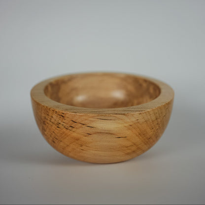 A small handcrafted wooden bowl with a natural light brown grain pattern, photographed against a neutral gray studio background.