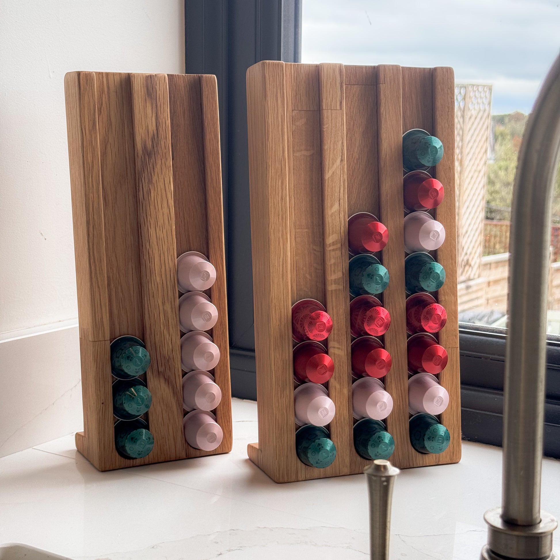 The large and small coffee pod holders on white worktop next to a grey window frame, the coffee pod stand are holding coffee capsules on colours red, pink and green and the shape of the coffee pod organiser is tall and rectangular