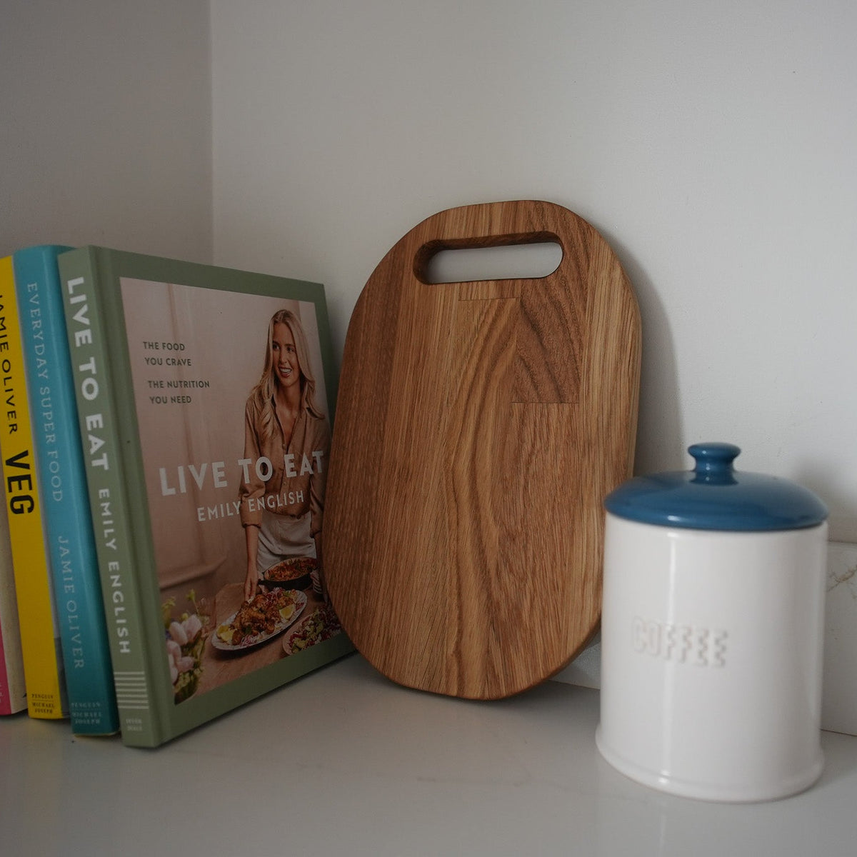 A wooden cutting board with a handle and a white ceramic canister with a blue lid sit on a white shelf next to a stack of cookbooks against a white wall.