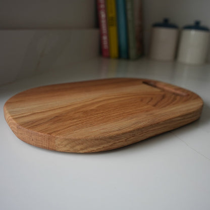 An oval-shaped wooden cutting board with a natural grain pattern and a small handle cutout, resting on a white countertop with a blurred kitchen background.