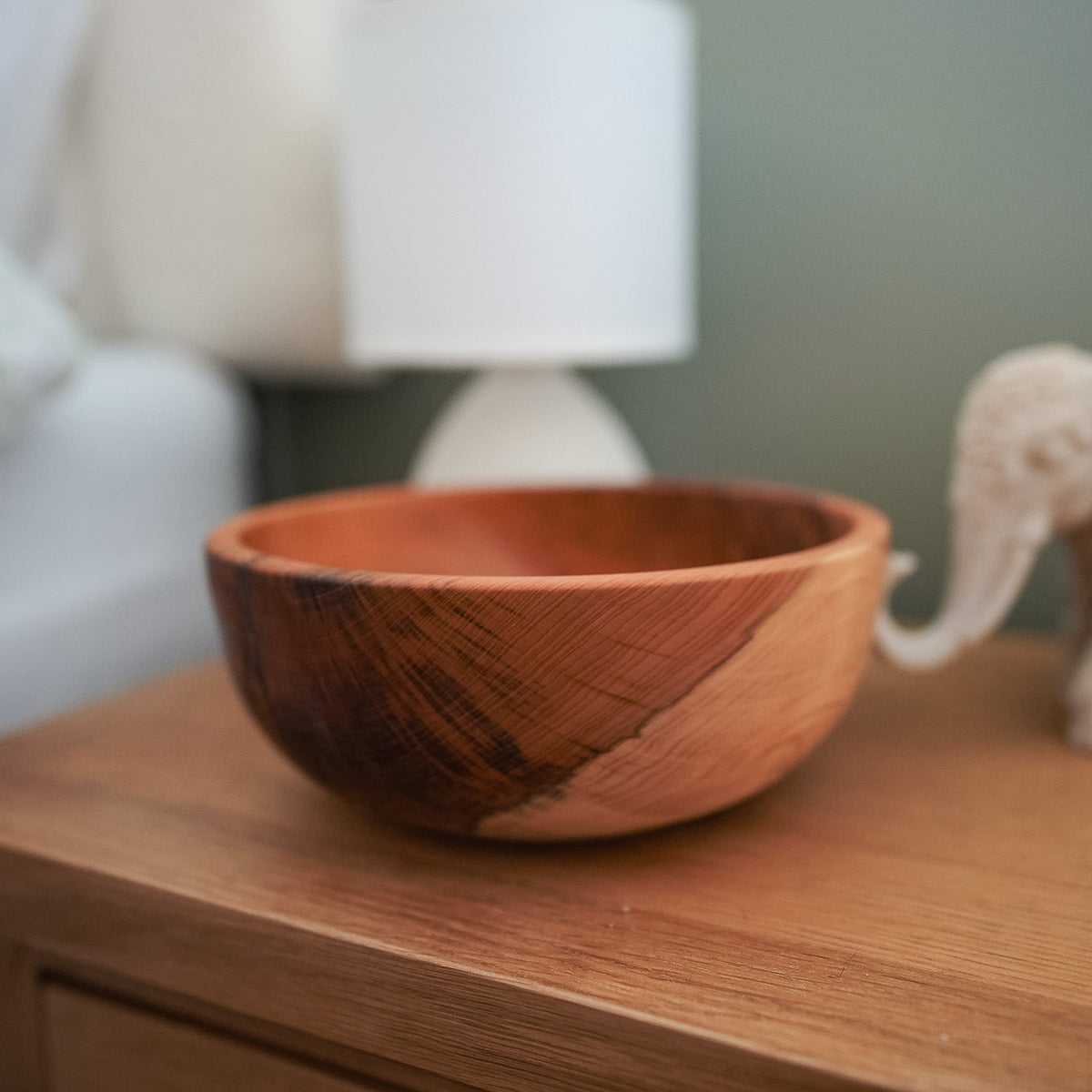A rustic wooden bowl with a dark brown and warm orange-brown wood grain pattern, sitting on a wooden bedside table next to a small elephant figurine and a blurred lamp in the background.