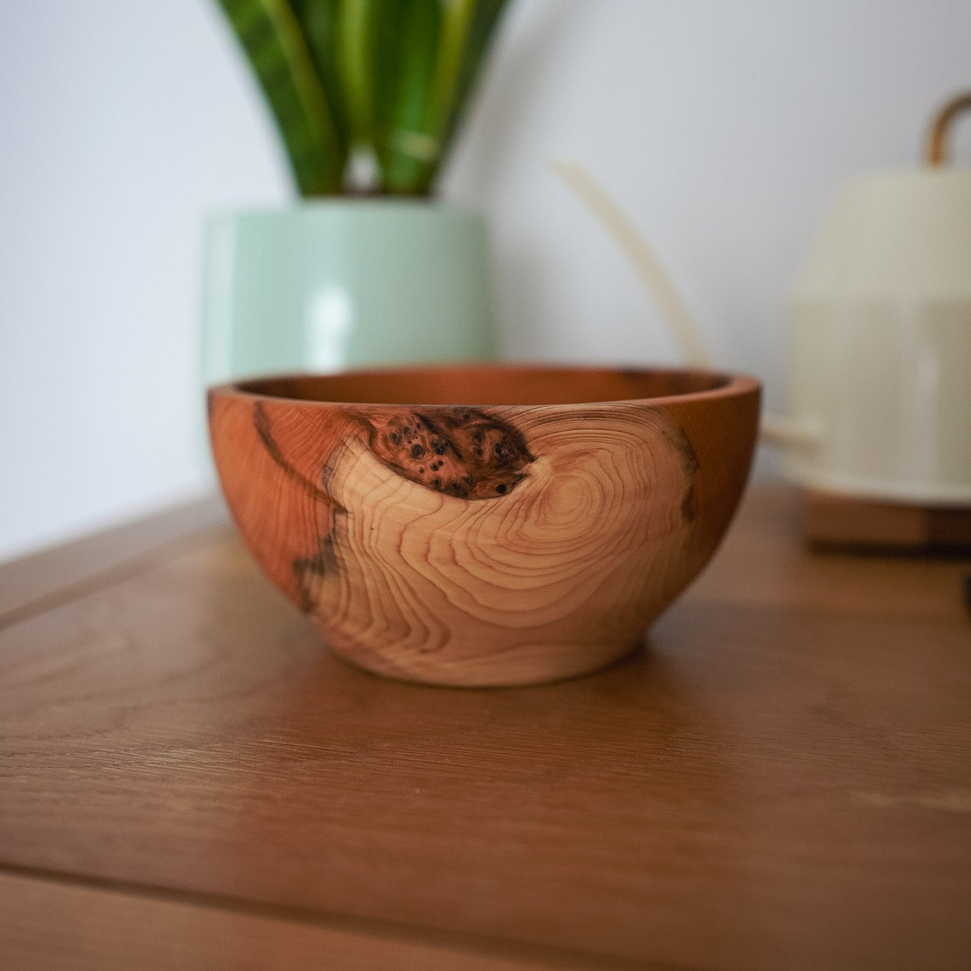 A handcrafted wooden bowl with a prominent natural knot and swirling wood grain, resting on a wooden table with a blurred potted snake plant in the background.