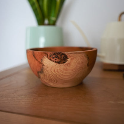 A handcrafted wooden bowl with a prominent natural knot and swirling wood grain, resting on a wooden table with a blurred potted snake plant in the background.