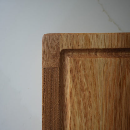 Close-up of a wooden cutting board with a thick raised border and visible wood grain, photographed against a light neutral wall.