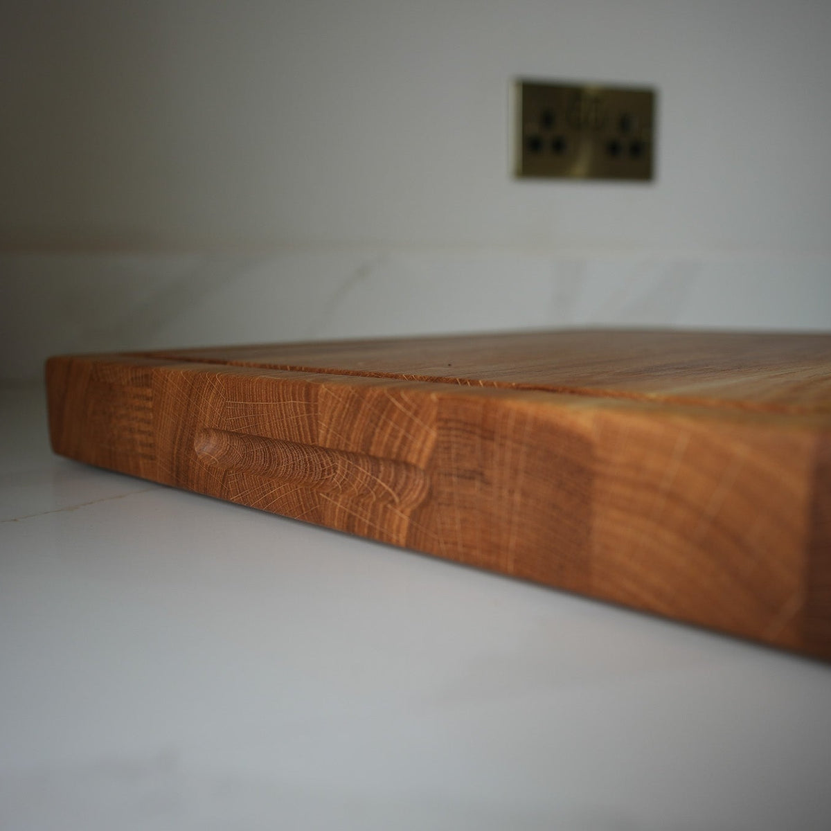 A thick, rectangular wooden cutting board with a warm honey-brown finish and visible wood grain, resting on a white marble countertop in a kitchen setting.
