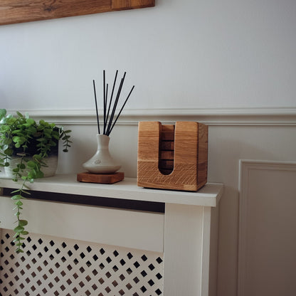 A wooden box with a cutout window for storing oak coasters styled on a white mantelpiece next to a potted green plant.