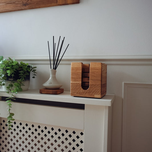 A wooden box with a cutout window for storing oak coasters styled on a white mantelpiece next to a potted green plant.