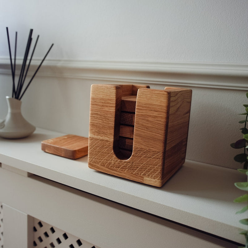 A rectangular wooden box organiser for coasters with a cutout front, shown on a white shelf next to a small matching wooden block and a diffuser, against a white wall.