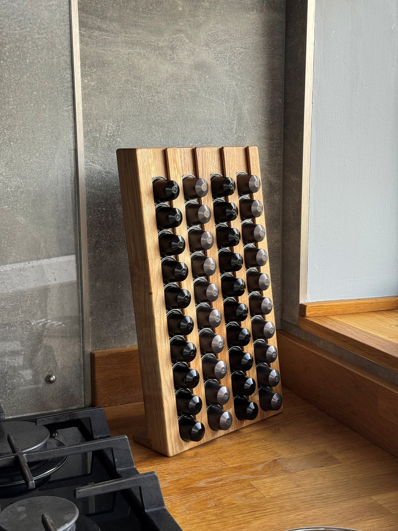 A wooden coffee pod organiser with multiple vertical slots set on a wooden kitchen counter against a gray backsplash.