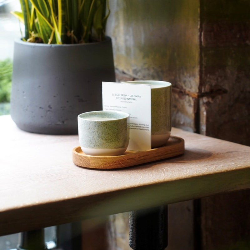 A small ceramic cup and matching candle in a wooden coffee tray with dipped rim sit on a wooden table beside a potted plant in a rustic indoor setting.