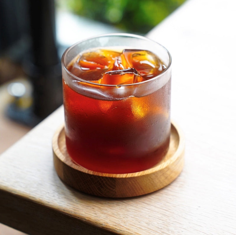 A frosted clear glass tumbler filled with iced iced coffee, resting on a round custom made wooden coaster with dipped rim on a light wood table.
