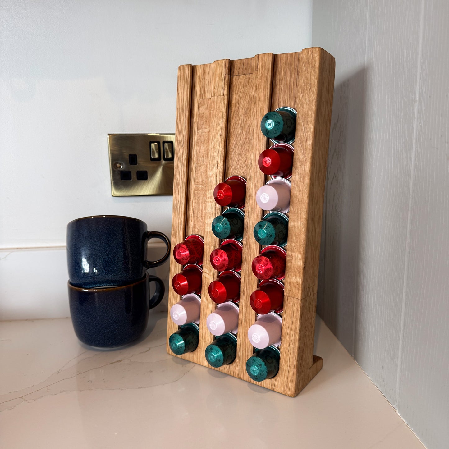 Large coffee pod stand with 27 green, pink and red coffee pods stacked, blue mugs next to it on a white counter with grey cupboard as background.