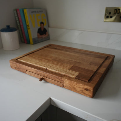 A rectangular wooden cutting board with a built-in drawer underneath, resting on a white marble kitchen countertop.