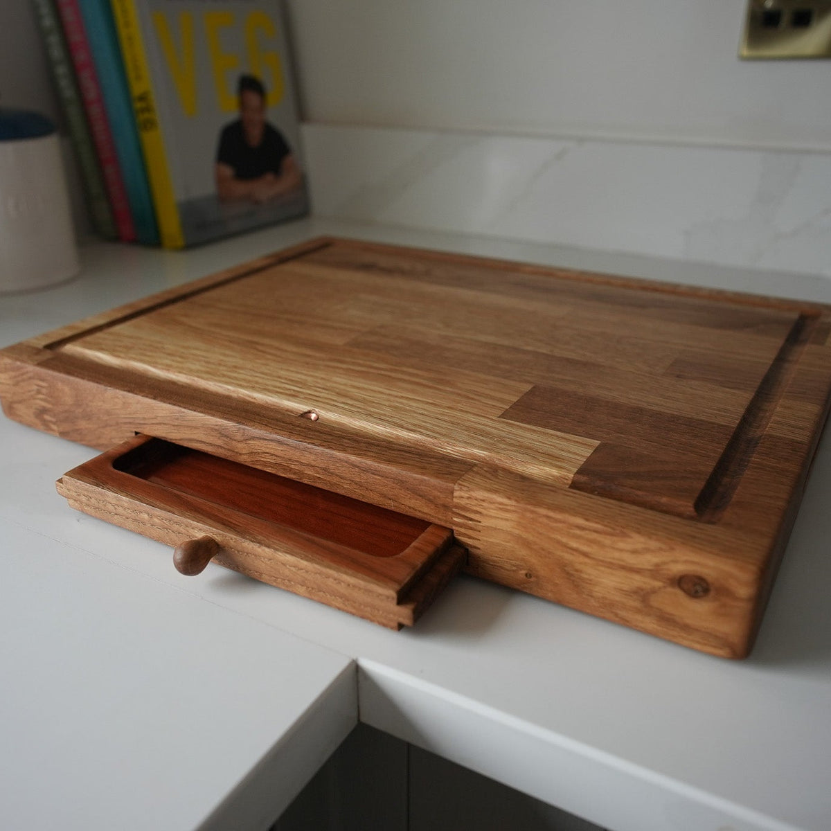 A rectangular wooden cutting board with a built-in drawer underneath, shown on a white kitchen countertop.